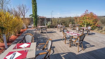 Un área de comedor al aire libre con mesas y sillas, rodeada de plantas y árboles.