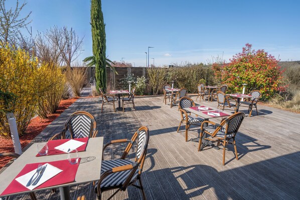 Un área de comedor al aire libre con mesas y sillas, rodeada de plantas y árboles.