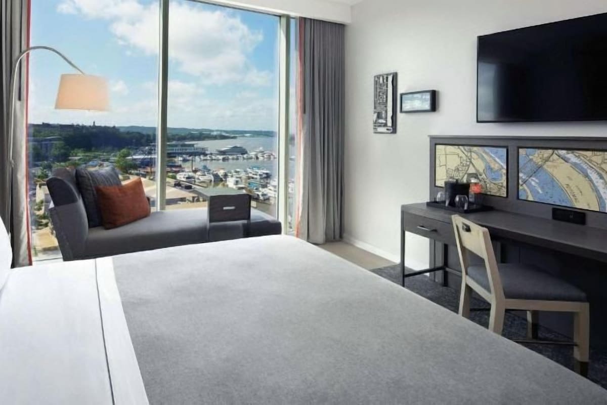 A hotel room with a large bed, a desk, a chair, a TV, and a view of a cityscape.