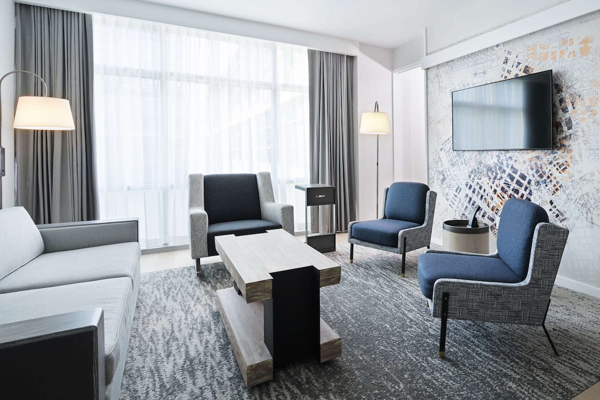 Moderni hotellihuone, jossa on sohva, kaksi nojatuolia, sohvapöytä, seinälle asennettu litteänäyttöinen televisio ja sängyssä pääty.