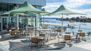 An outdoor terrace with tables and chairs, large umbrellas, and a view of a body of water.