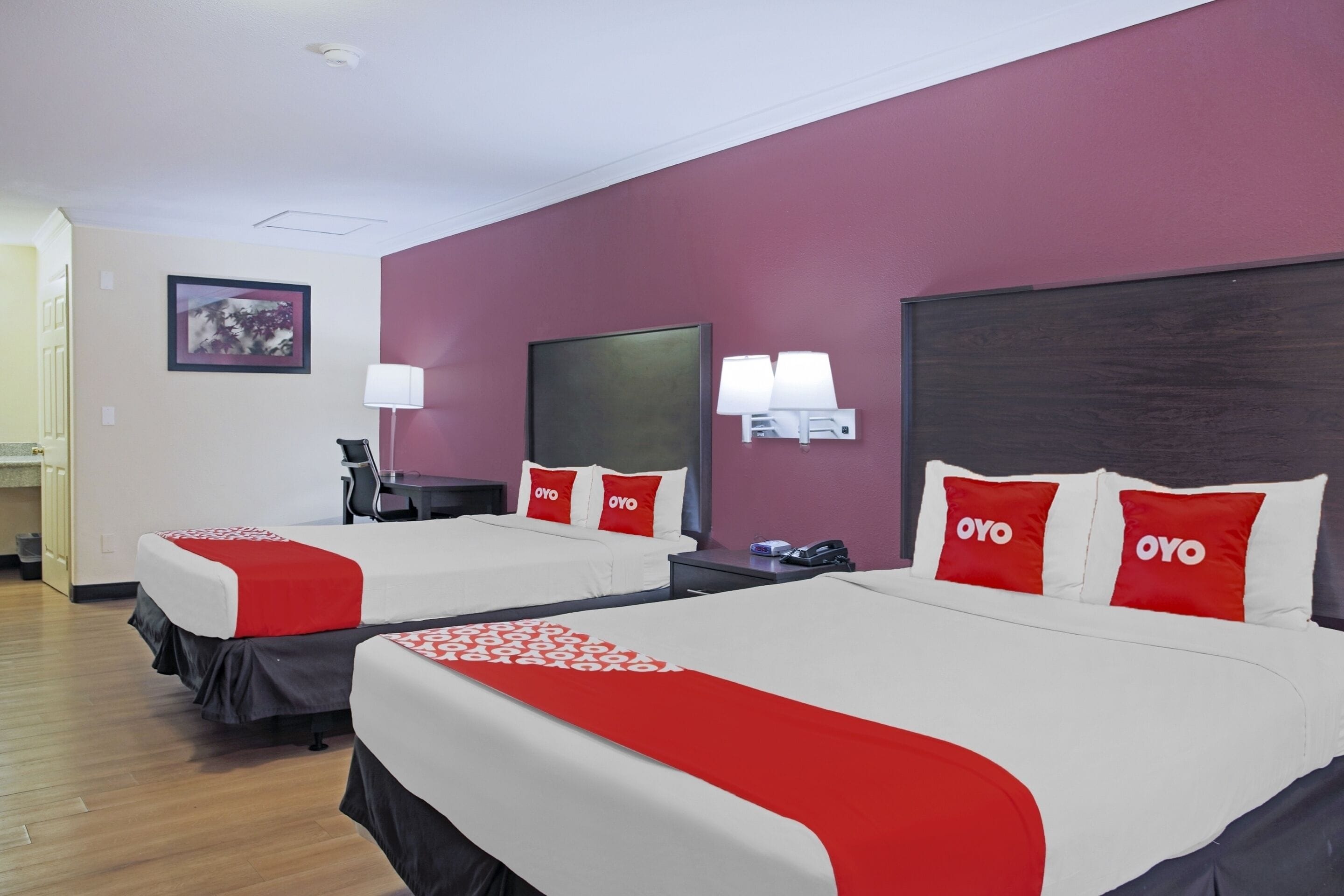 A hotel room with two beds, each with red and white bedding and pillows labeled "OYO". There are wall-mounted lamps and a framed picture on the wall.