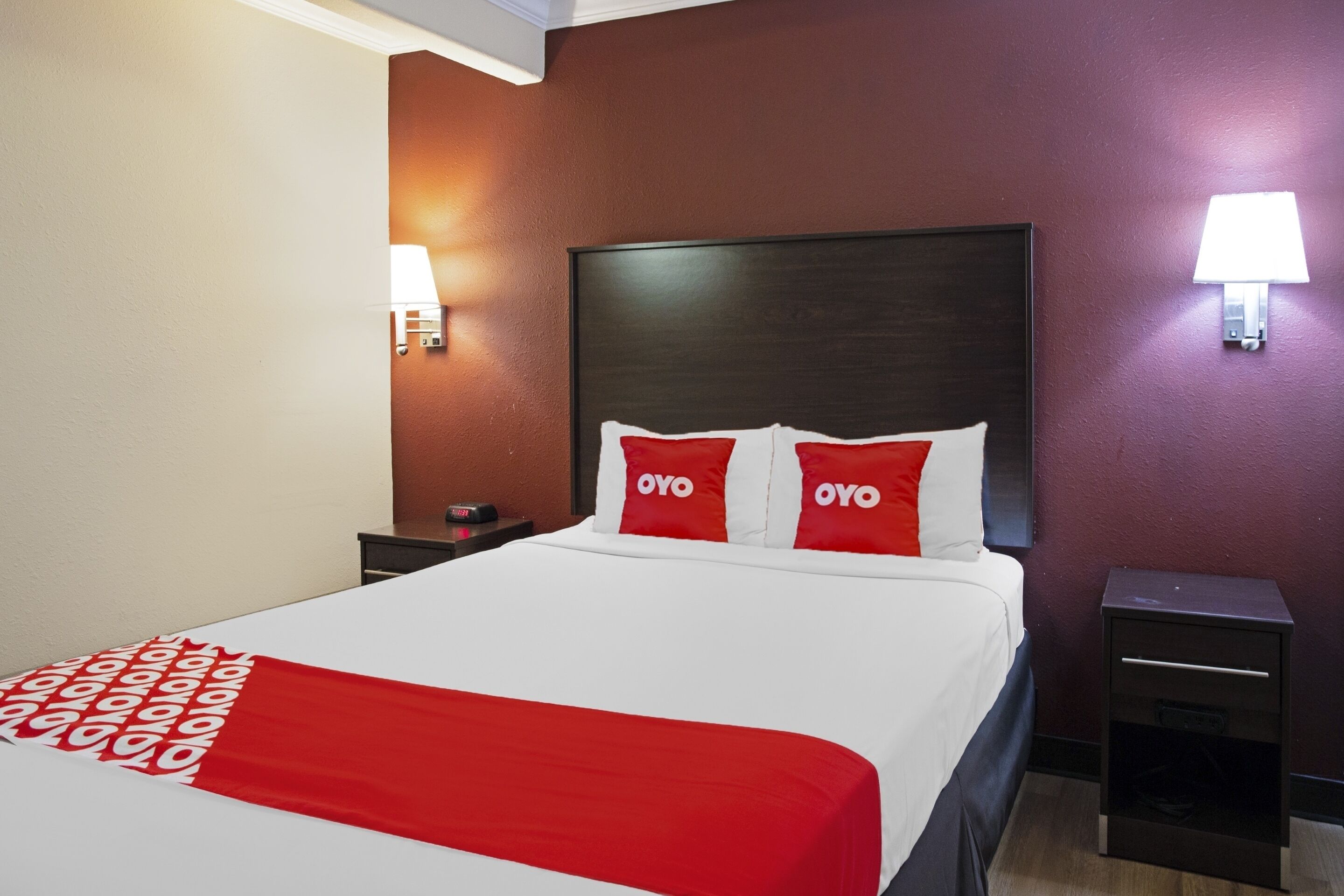 A hotel room with a bed, two bedside tables, a nightstand, a red and white patterned throw blanket, and two wall-mounted lamps.