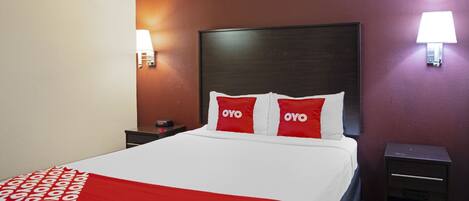 A hotel room with a bed, two bedside tables, a nightstand, a red and white patterned throw blanket, and two wall-mounted lamps.