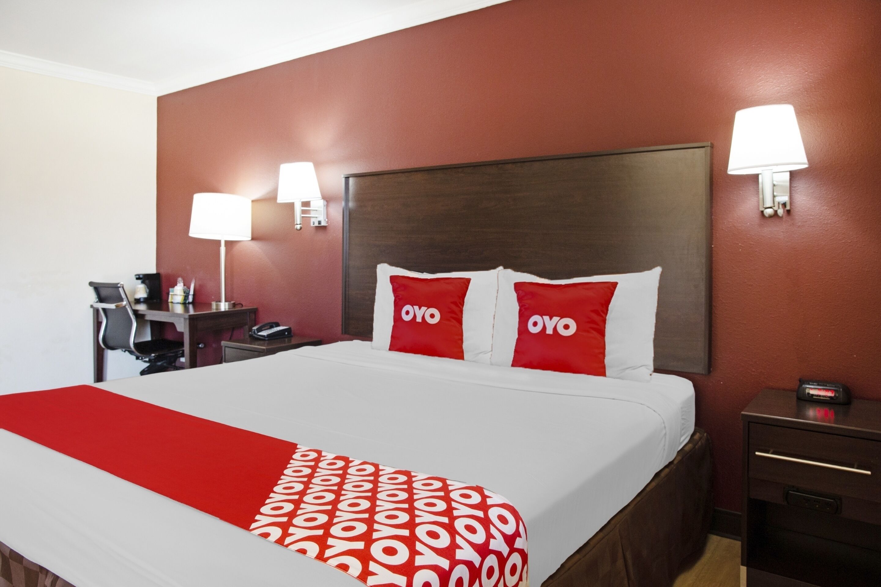 A hotel room with a large bed, two bedside lamps, a desk with a chair, and a nightstand with a clock.