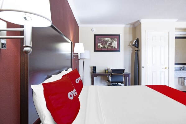 Habitación de hotel con cama, escritorio, silla, lámpara, un cuadro en la pared y una puerta.