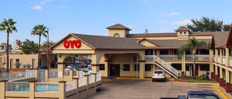 Un hotel con un cartel llamativo de 'OYO', un estacionamiento con varios coches y una zona de piscina con valla.