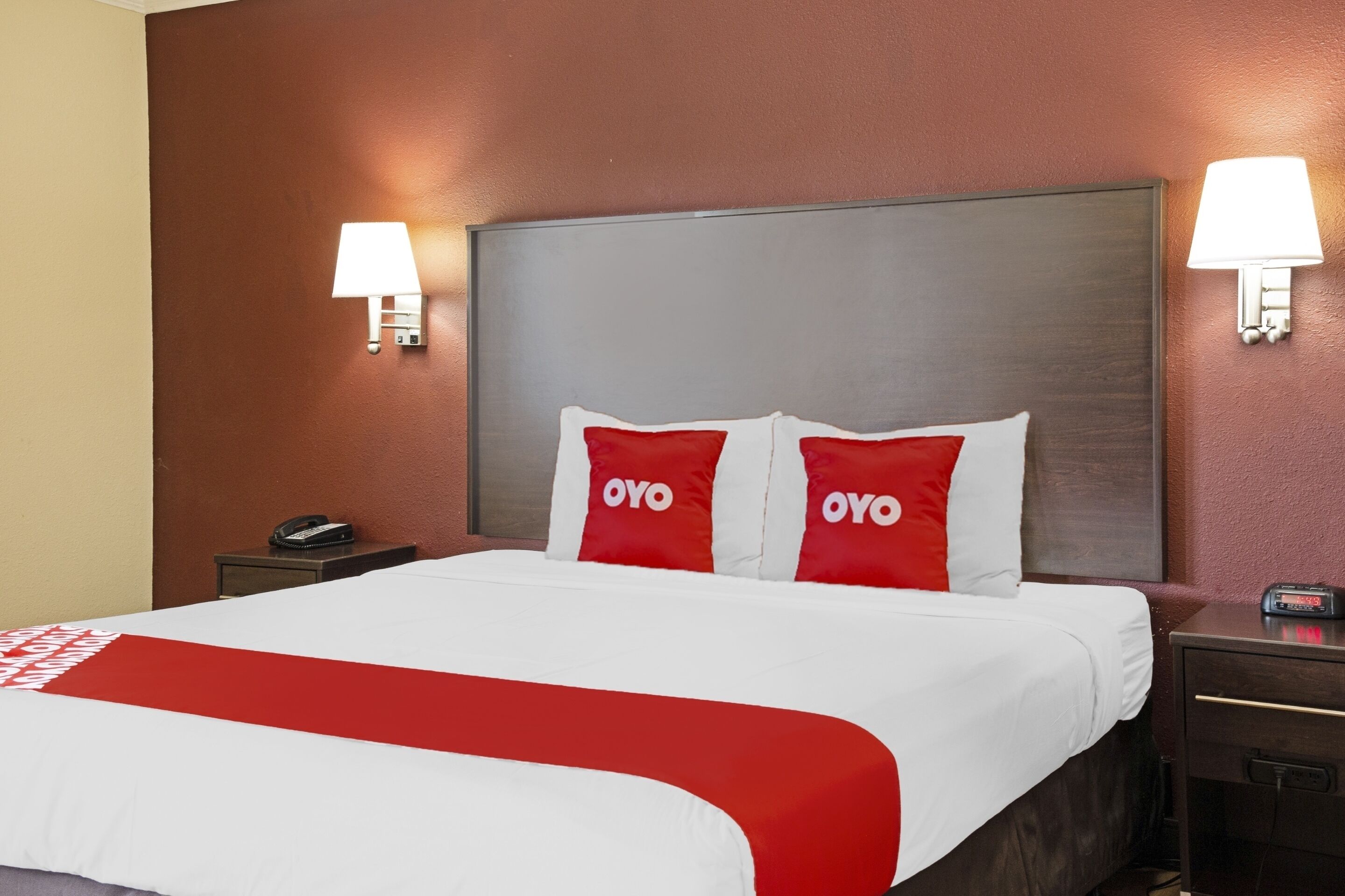 A hotel room with a large bed, two bedside lamps, a nightstand with a phone, and a digital clock.