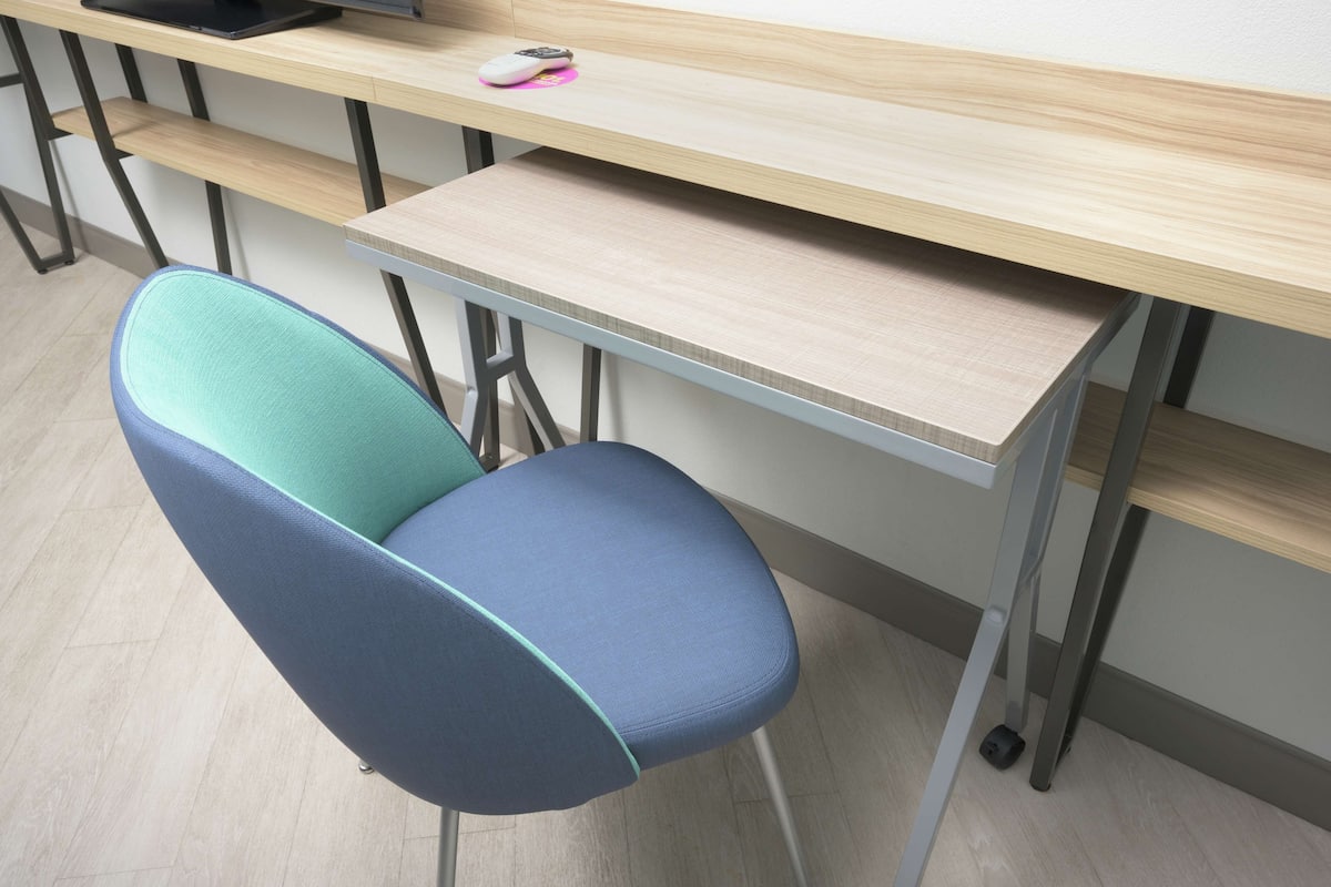 A modern office chair with a teal and blue backrest, positioned in front of a wooden desk with a pink mouse pad and a remote control on it.