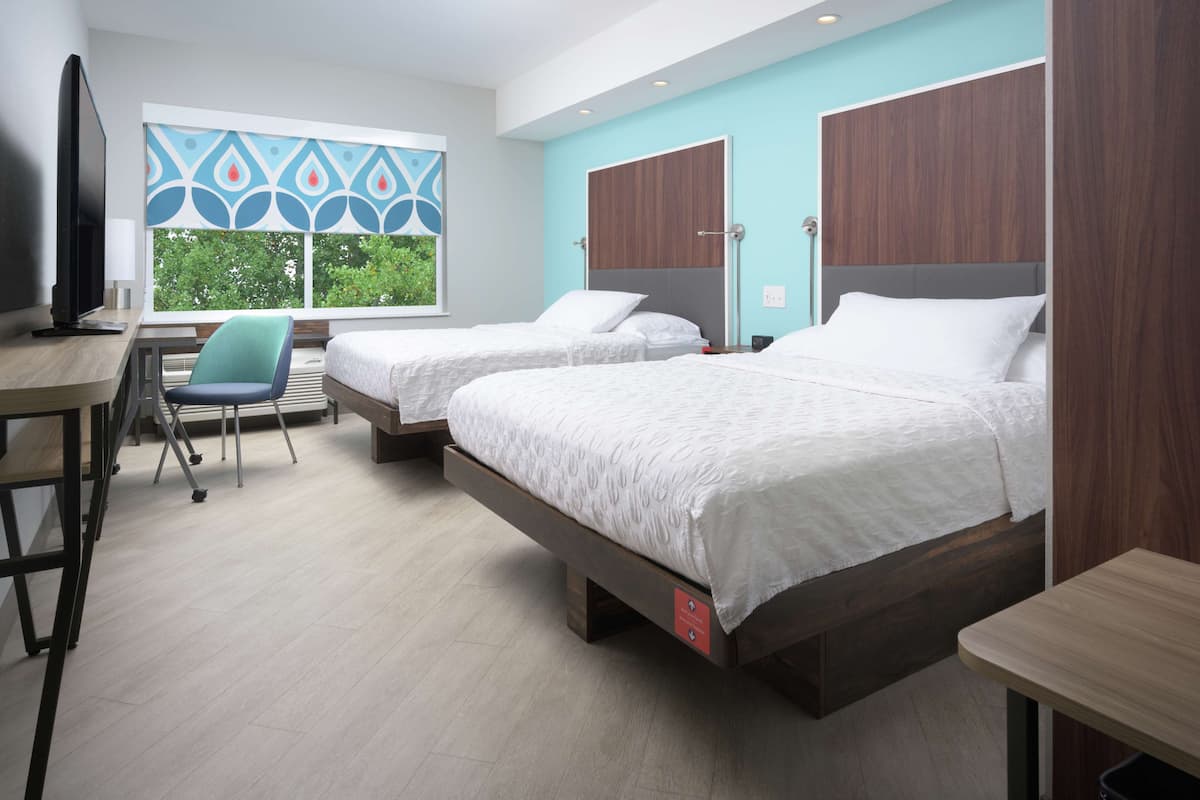A hotel room with two beds, a desk, a chair, and a TV.