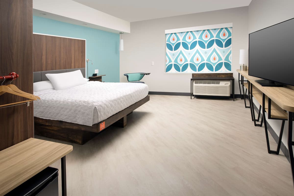 A modern hotel room with a bed, a desk, a chair, a TV, and a large window with a patterned view.