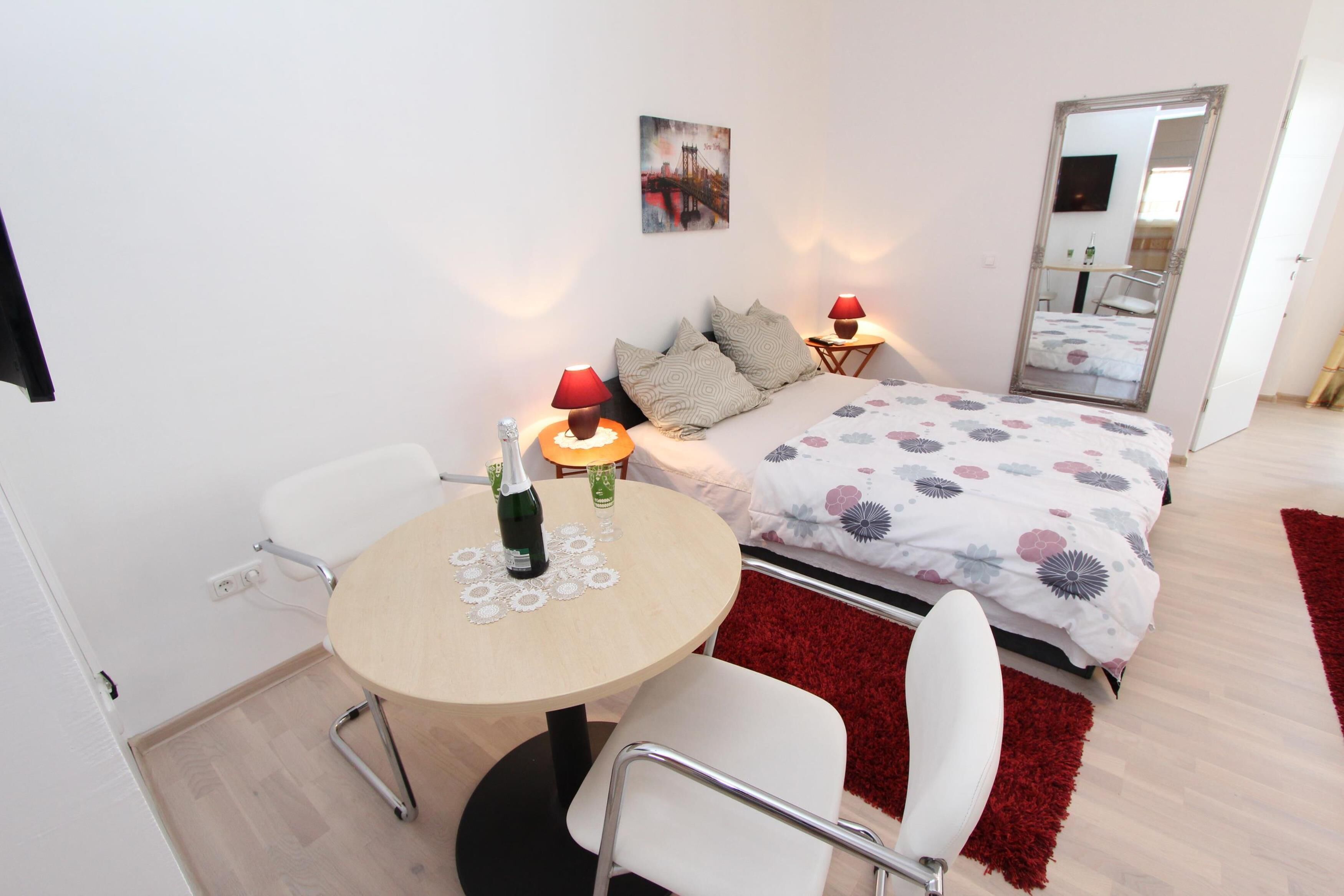 A bedroom with a bed, bedside tables, a round table with chairs, a red rug, and a wall-mounted picture.