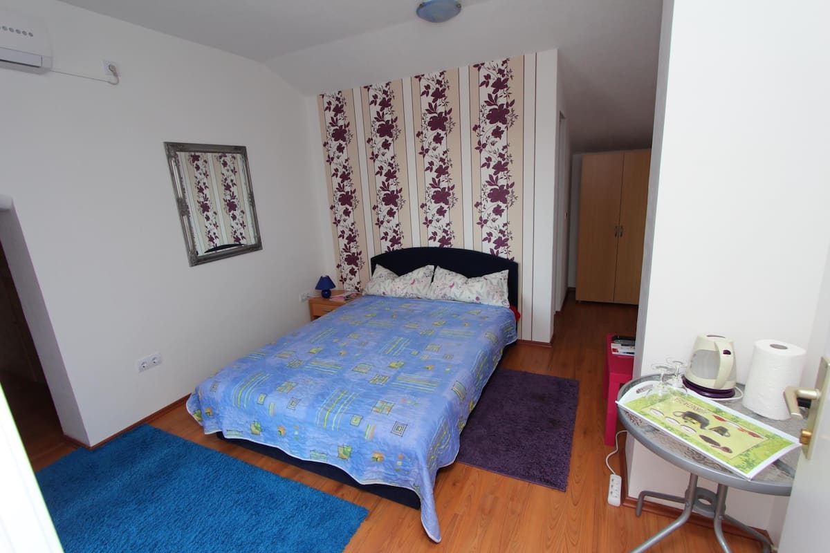 Um quarto pequeno com uma cama, mesa de cabeceira, janela com cortinas florais, porta e uma pequena mesa com chaleira e rolo de papel higiênico.