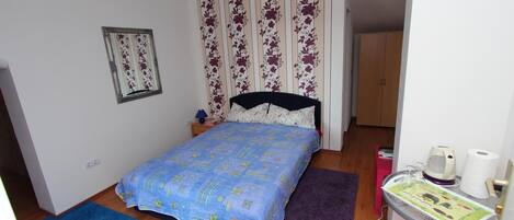 A small bedroom with a bed, a nightstand, a window with floral curtains, a door, and a small table with a kettle and a roll of toilet paper.