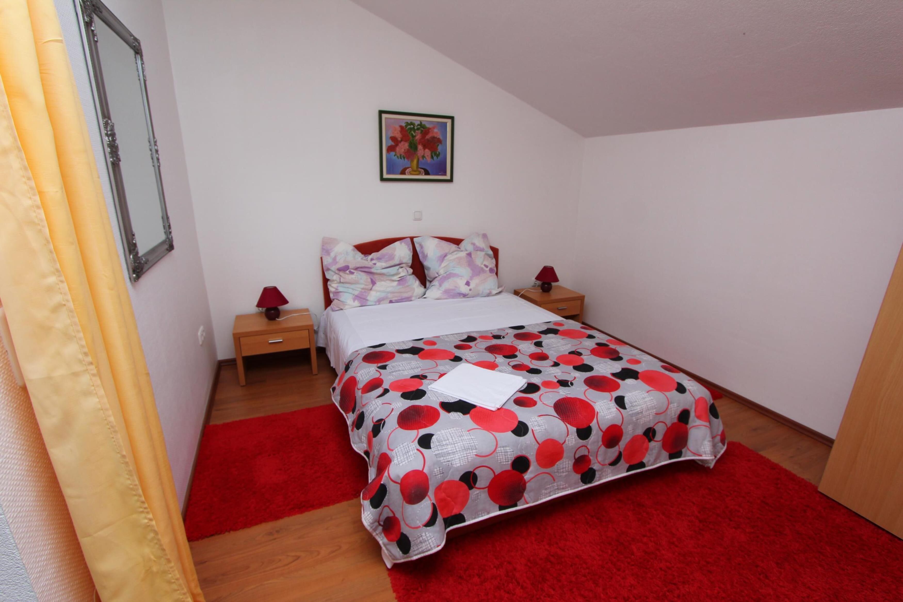 Un dormitorio con cama, mesita de noche, alfombra roja y un cuadro en la pared.