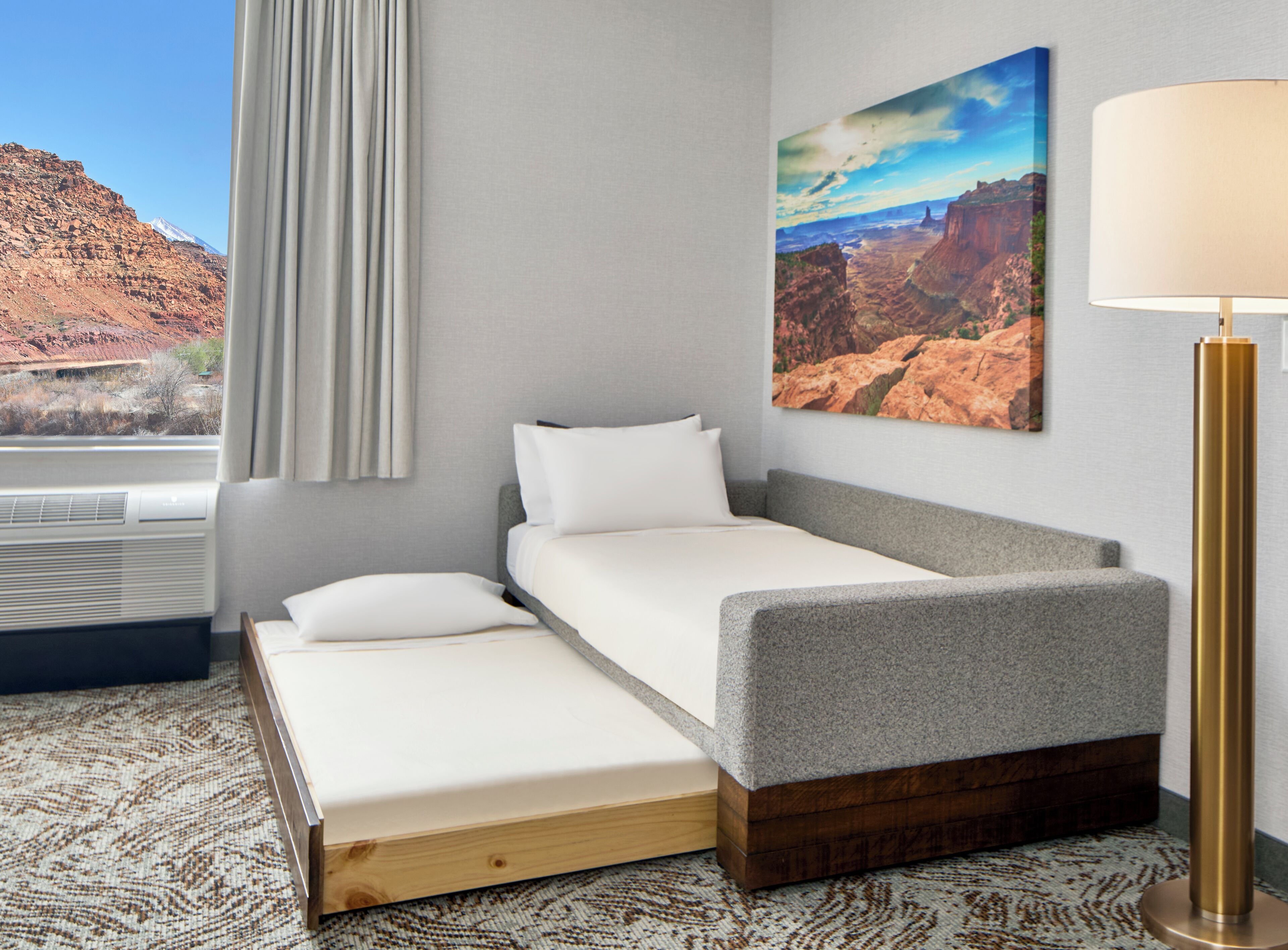Habitación de hotel con cama, mesita de noche, lámpara, un cuadro y ventana con cortinas.