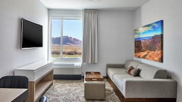 A modern hotel room with a flat-screen TV, a sofa, a desk, and a large window offering a view of the desert landscape.