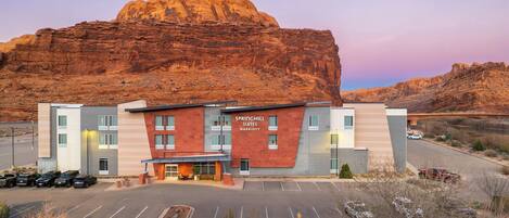 A hotel with a modern design, a parking lot, and a large rock formation in the background.