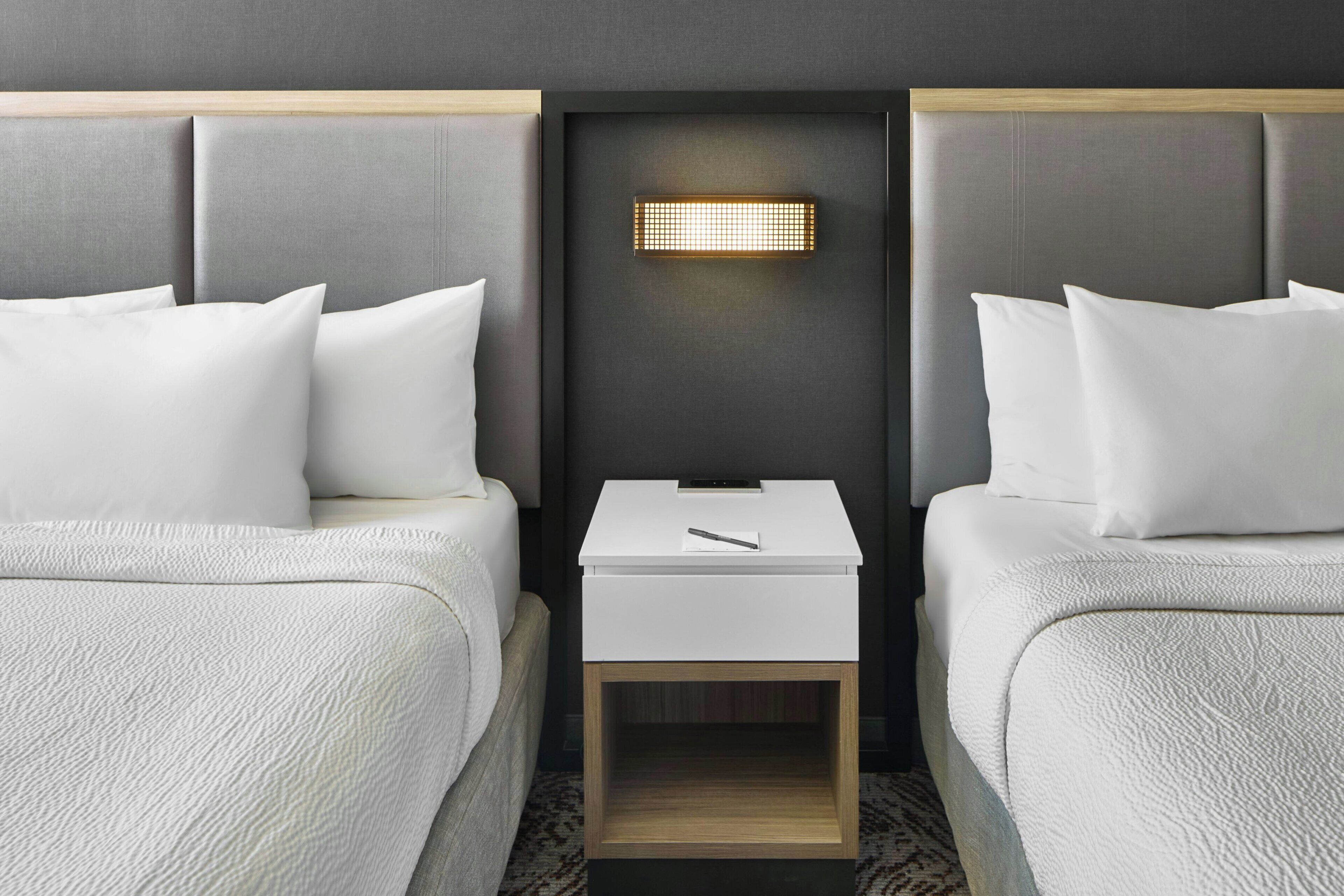 A hotel room with two beds, a bedside table, and a wall-mounted light fixture.