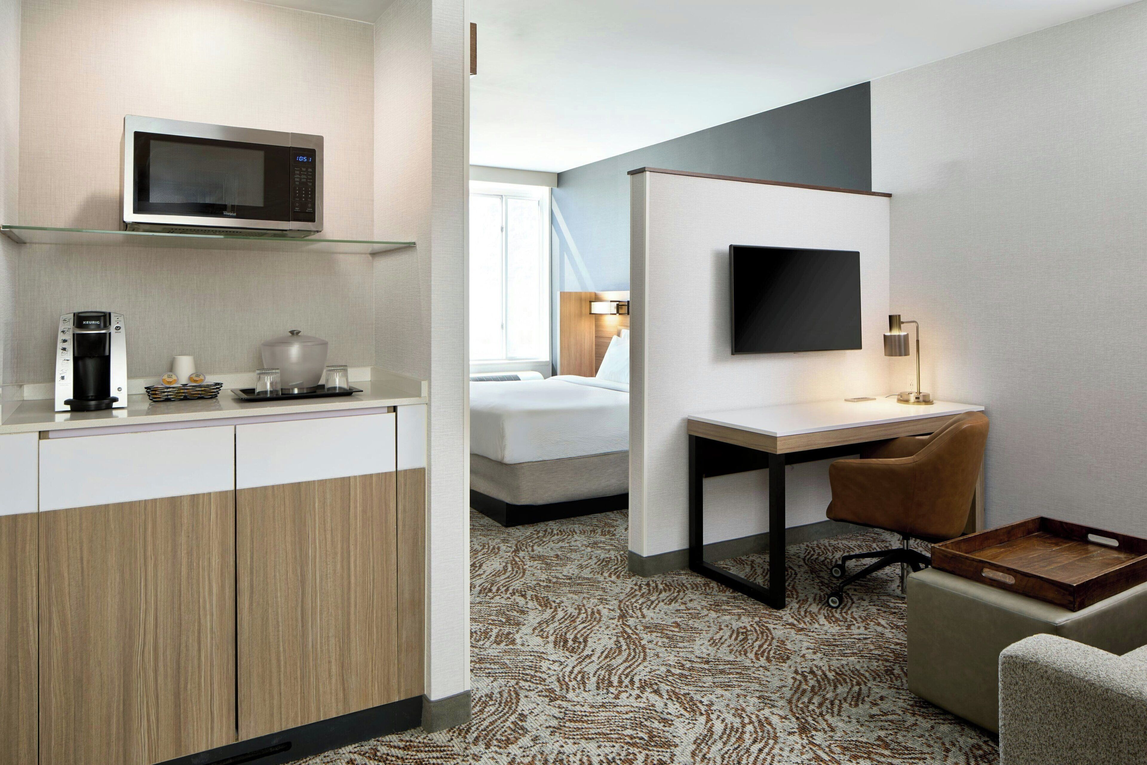 A hotel room with a kitchenette, a bed, a desk with a chair, a TV, and a patterned carpet.