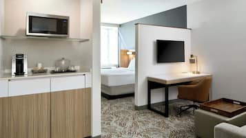 A hotel room with a kitchenette, a bed, a desk with a chair, a TV, and a patterned carpet.