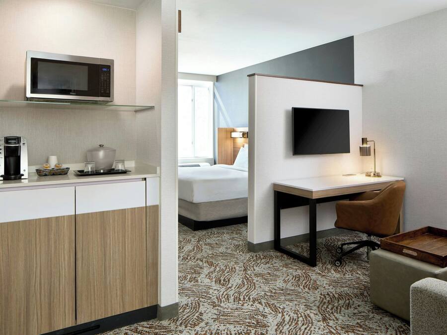A hotel room with a kitchenette, a bed, a desk with a chair, a TV, and a patterned carpet.