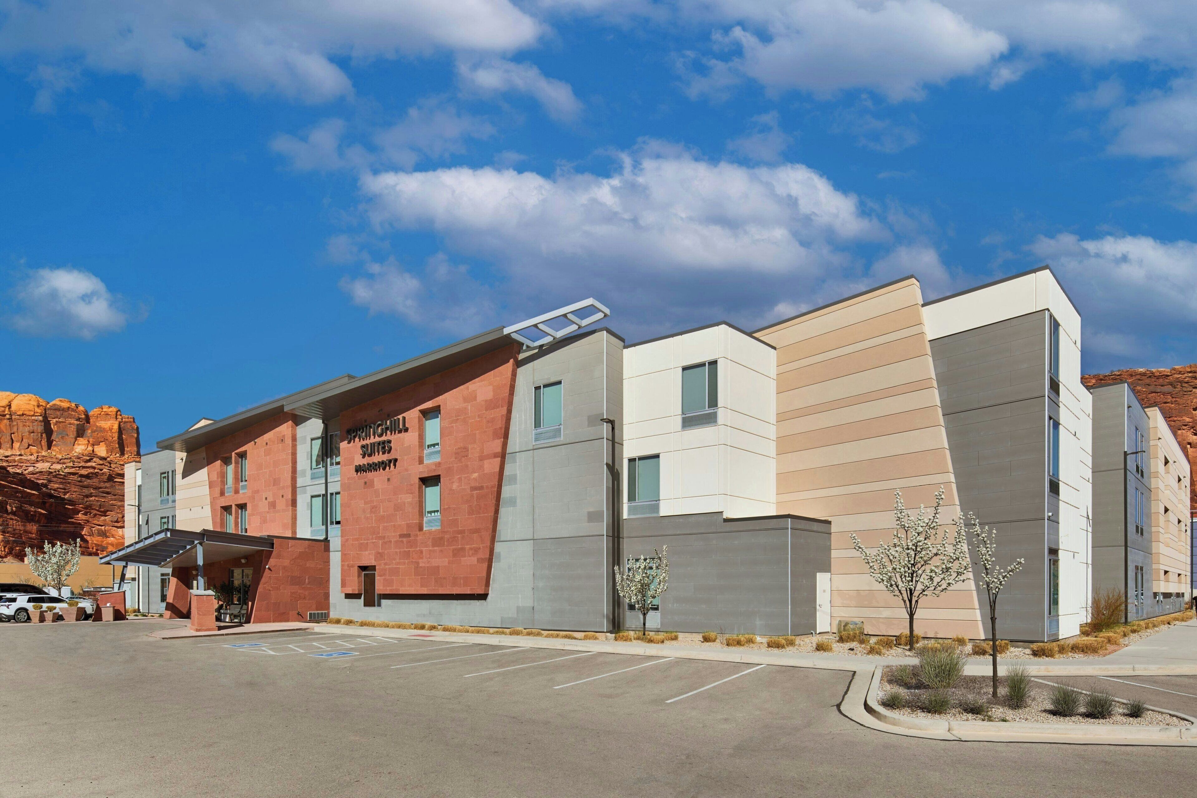 A modern hotel with a flat roof, a parking lot, and a desert landscape in the background.