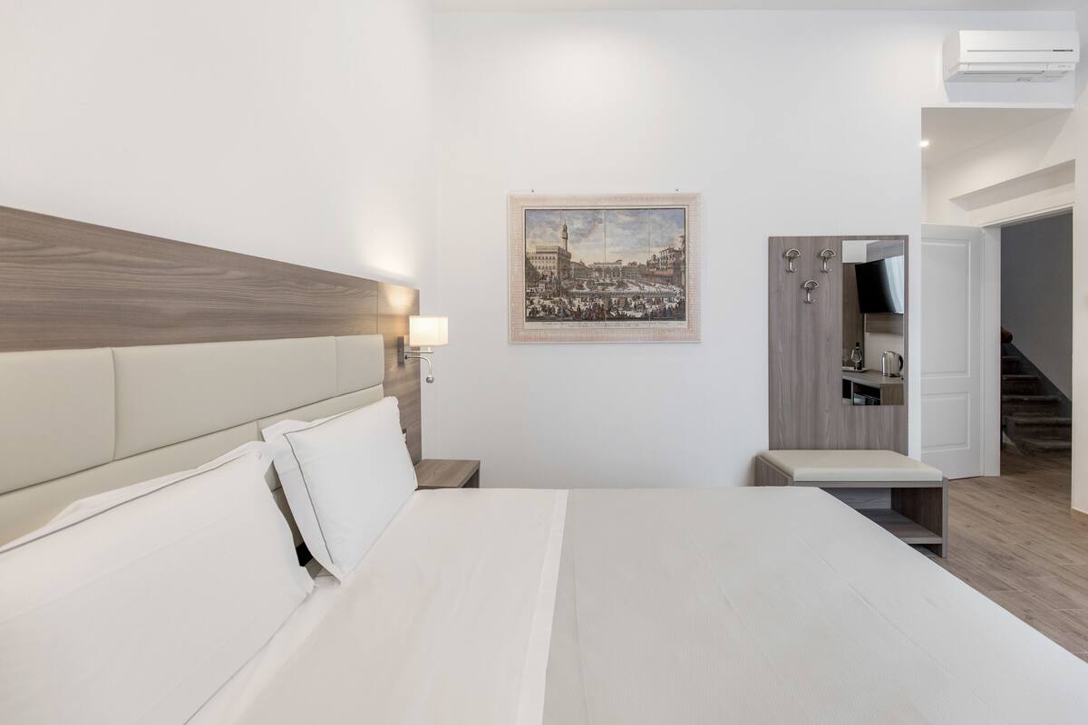Une chambre d’hôtel moderne avec un grand lit, une table de chevet, une lampe fixée au mur, un tableau représentant un paysage urbain, un petit banc et une porte donnant sur la salle de bain.