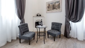 A room with two grey upholstered armchairs, a small round table with a wine glass and a book, a fireplace with a framed painting above it, and a white wall.