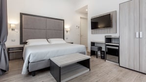 A modern hotel room with a large bed, bedside tables, a TV, a wardrobe, and a desk with a chair.