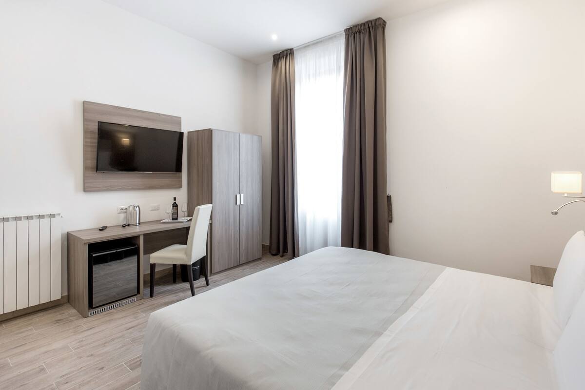 Une chambre d’hôtel comprenant un lit, un bureau avec une chaise, une télévision, une armoire et une fenêtre avec des rideaux.