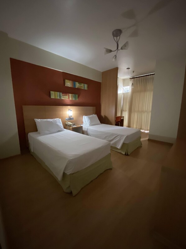 A hotel room with two beds, a ceiling fan, a bedside table with a lamp, and a window with curtains.
