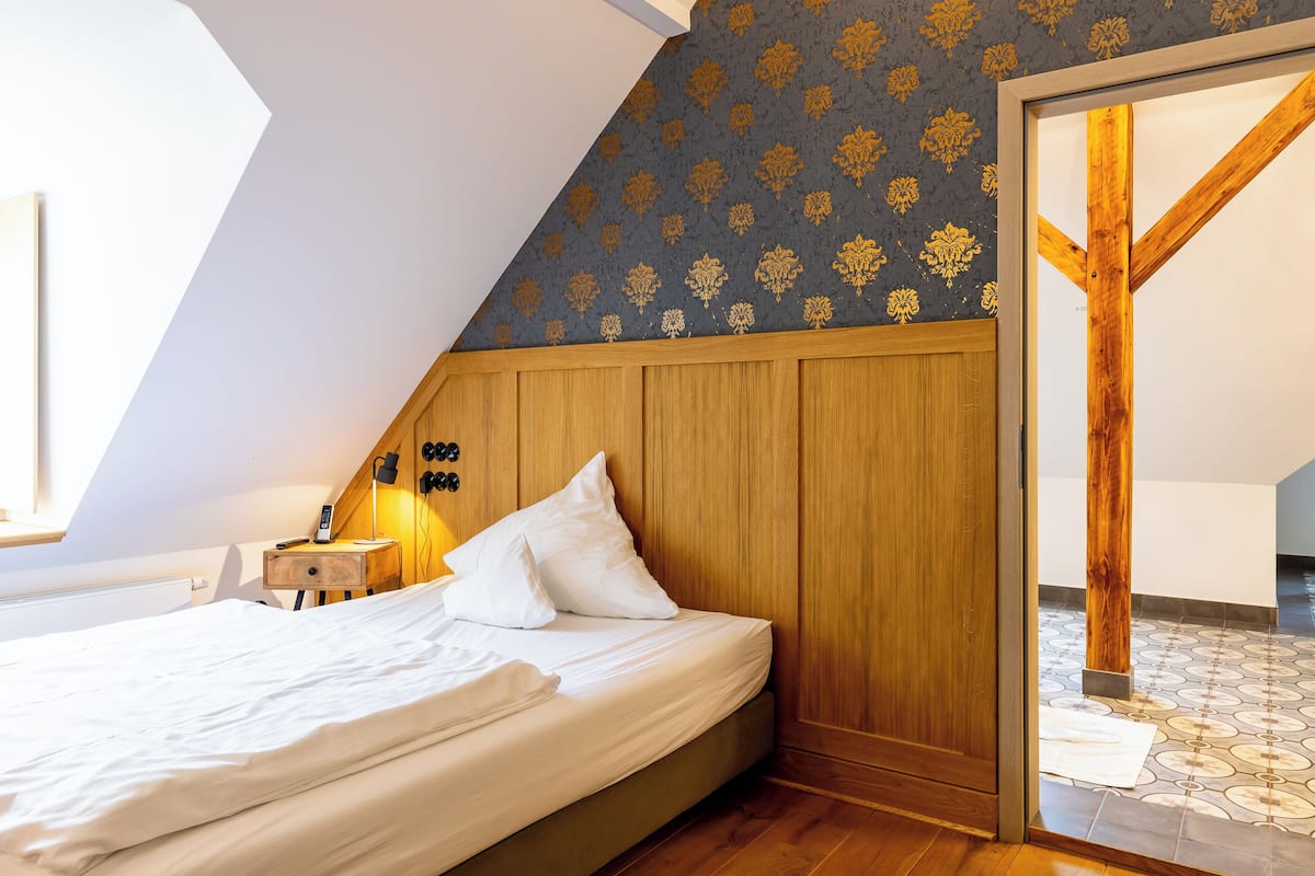 A bedroom with a sloped ceiling, wooden headboard, and a patterned wallpaper.