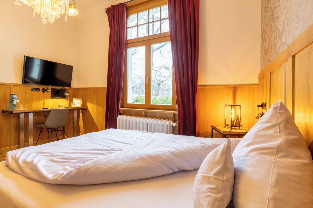 A hotel room with a bed, a desk with a chair, a television, and a window with curtains.