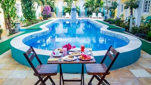 Una piscina con una fuente, rodeada por un patio empedrado con dos sillas de madera y una mesa lista para el desayuno.