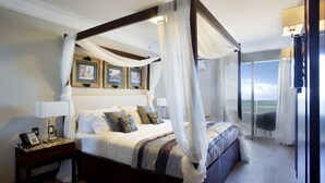 A four-poster bed with a canopy, a nightstand with a lamp, framed pictures on the wall, and a view of the outdoors through a large window. - Royal Atlântica Macaé Hotel (Macaé)