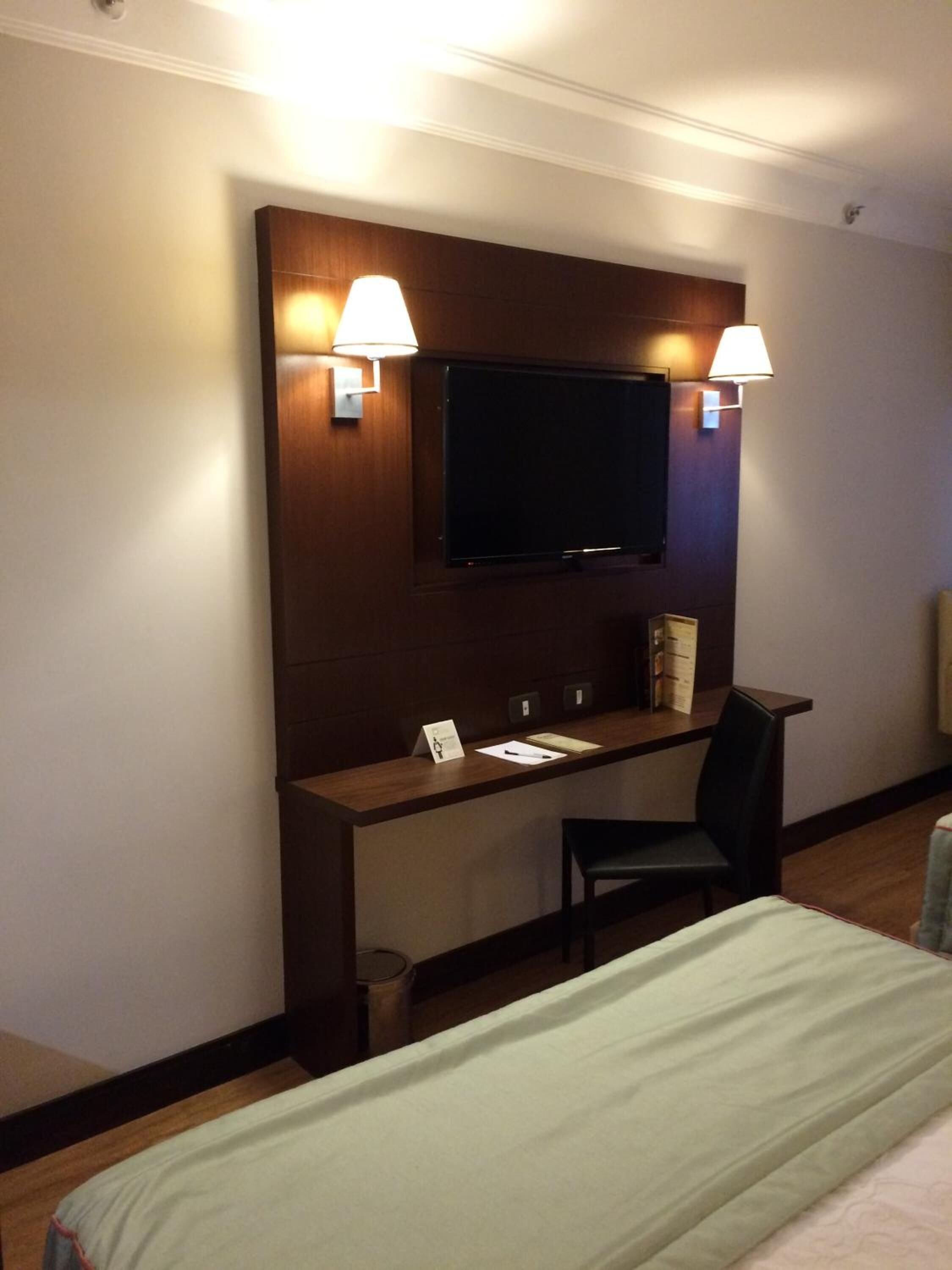 A hotel room with a bed, a desk with a chair, a television mounted on the wall, and two wall-mounted lamps.