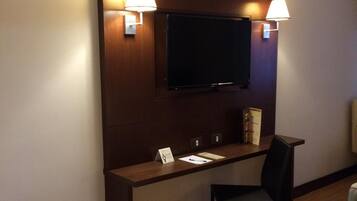 A hotel room with a bed, a desk with a chair, a television mounted on the wall, and two wall-mounted lamps.