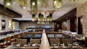 A modern restaurant with long communal tables, hanging plants, and a stone wall. - Royal Atlântica Macaé Hotel (Macaé)