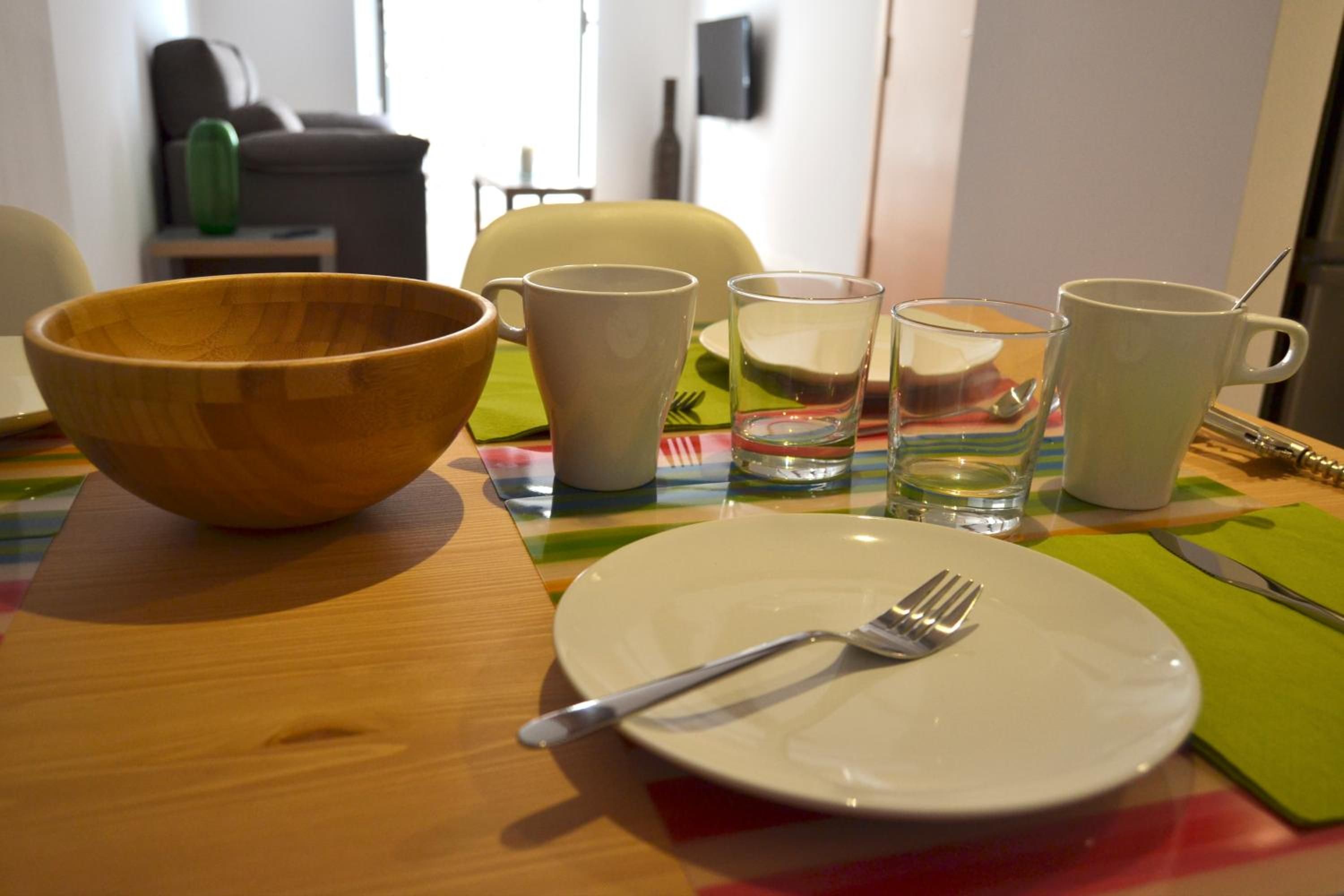 Une table à manger avec un saladier en bois, une assiette blanche avec une fourchette, ainsi que plusieurs tasses et verres blancs.