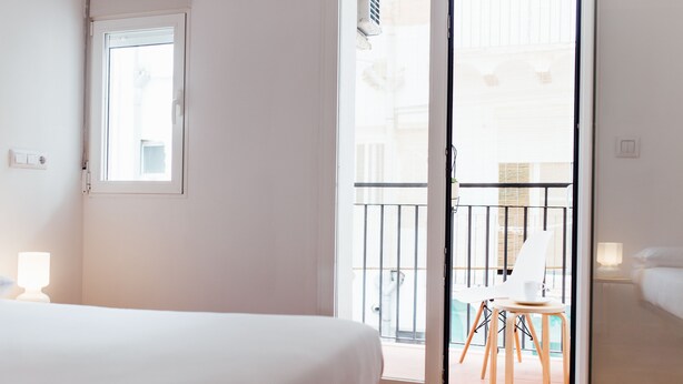 Habitación de hotel con cama, ventana y puerta corrediza de vidrio que da a un balcón exterior con una silla y una mesa.