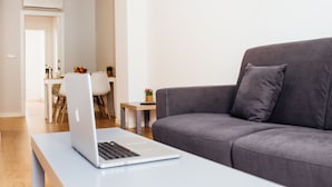Una sala de estar moderna con un sofá gris, una computadora portátil sobre una mesa blanca y una mesita auxiliar con una planta.