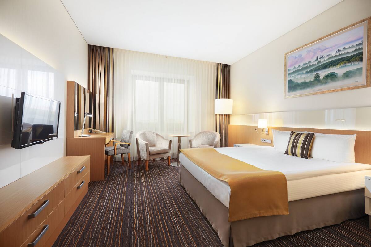A hotel room with a bed, a desk with a chair, a television, and a painting on the wall.