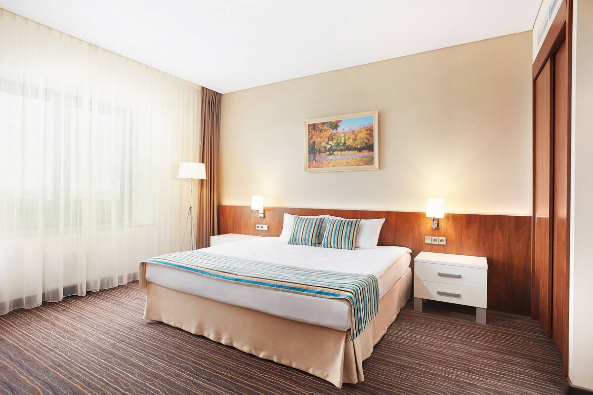 A hotel room with a large bed, two bedside tables, a nightstand, a wall-mounted lamp, and a painting.