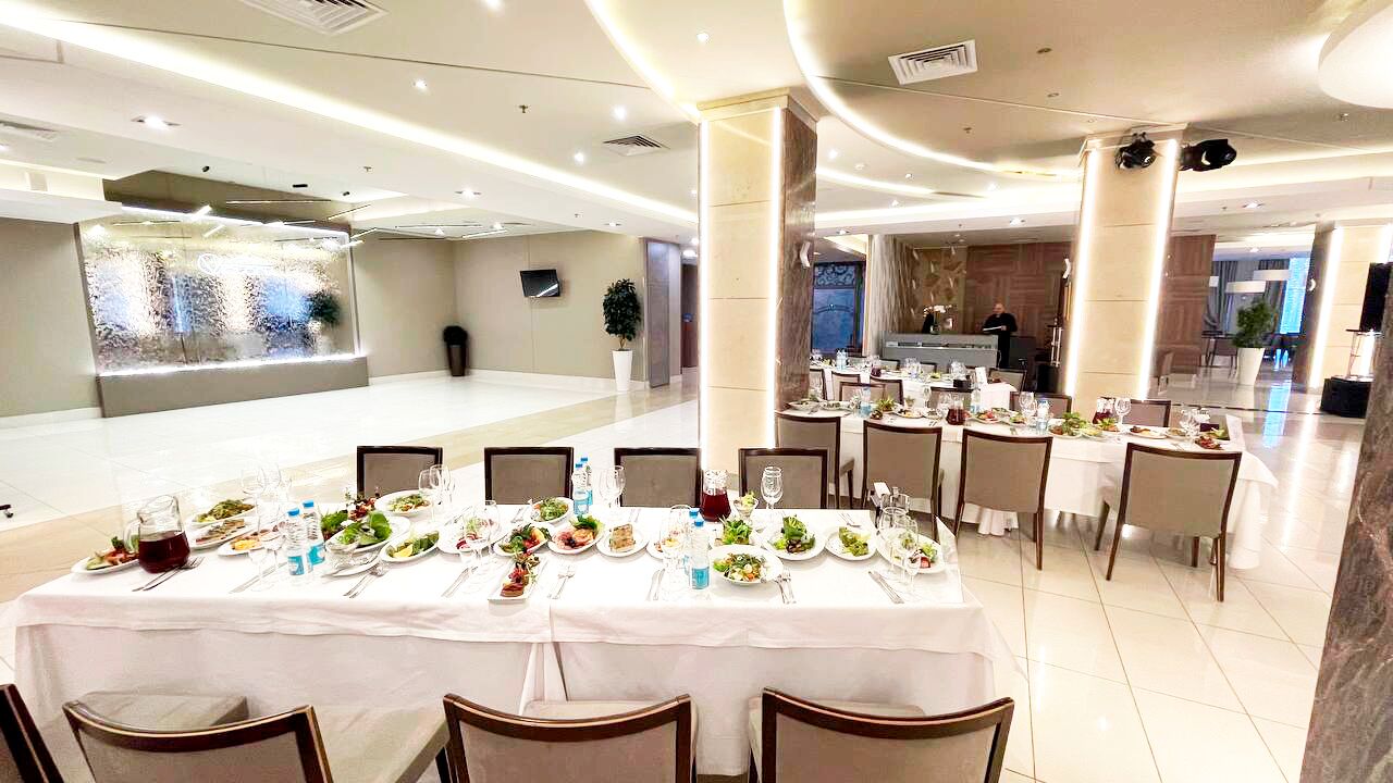 Un vaste espace dédié à la restauration, avec plusieurs tables dressées pour les repas, un comptoir d’accueil et un aménagement intérieur moderne.