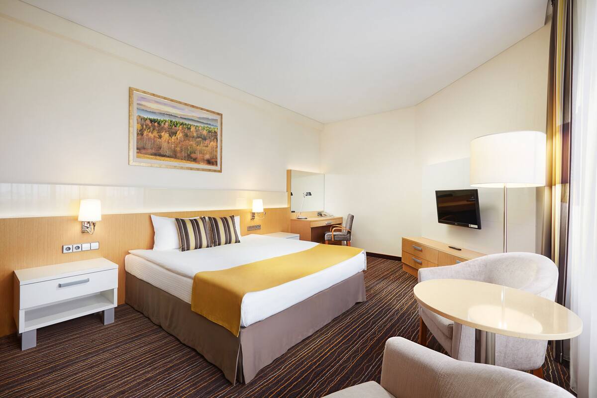 A hotel room with a large bed, bedside tables, a desk, a chair, a TV, and a painting on the wall.