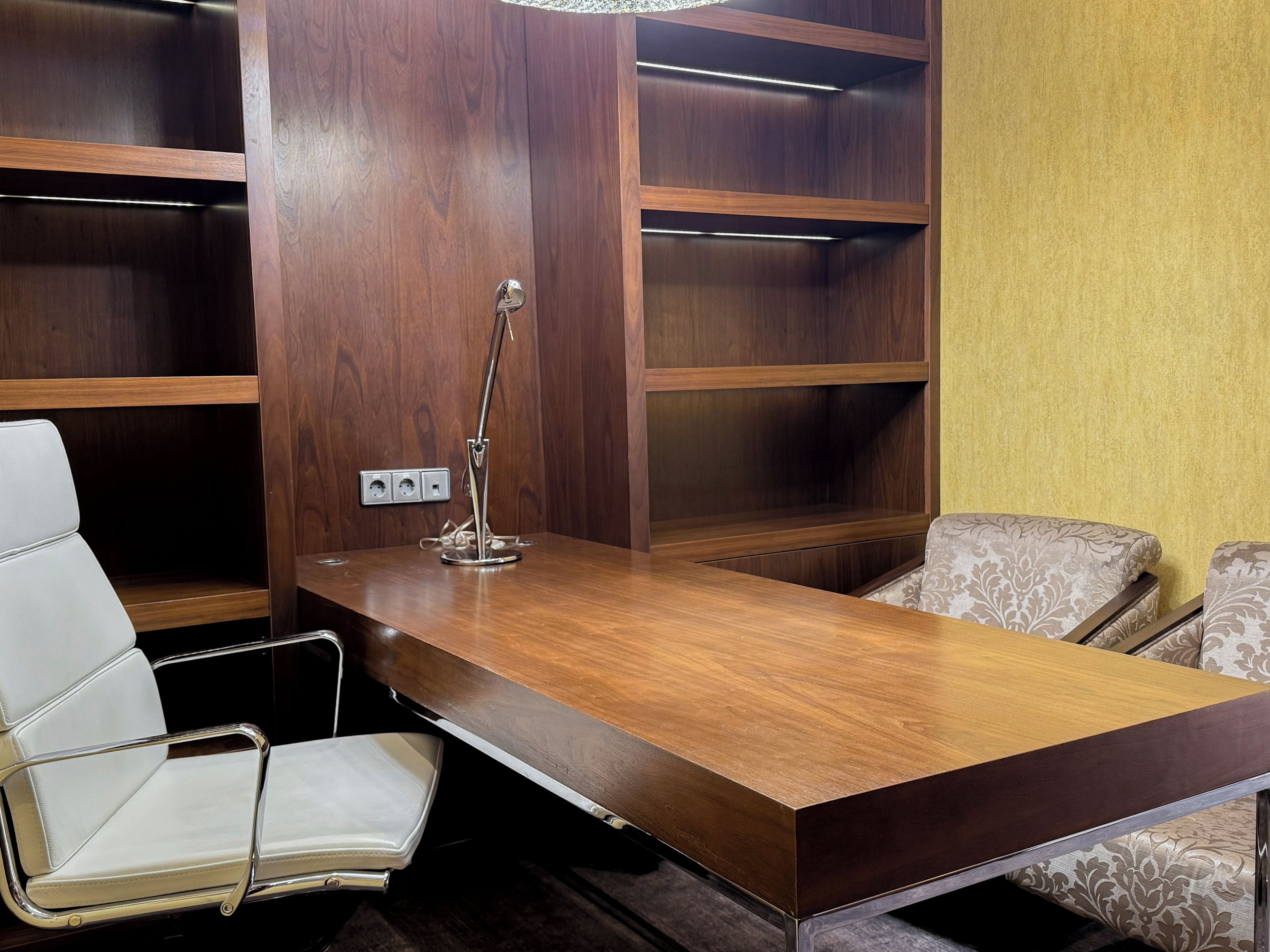 A conference room with a wooden table, two chairs, a desk lamp, and built-in shelving.