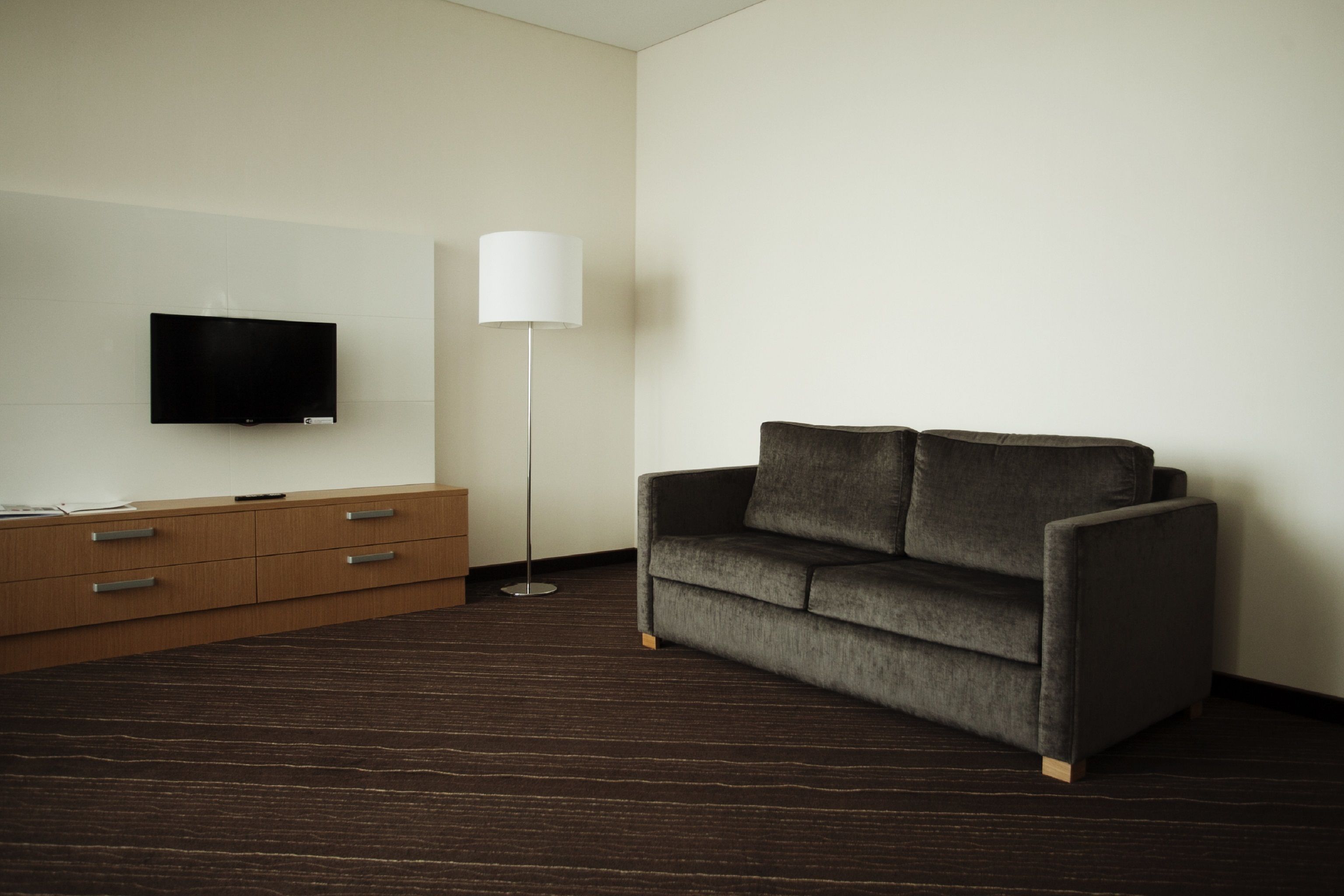 Una habitación de hotel moderna con piso de madera oscura, un sofá gris, un televisor de pantalla plana montado en la pared y una lámpara de pie.