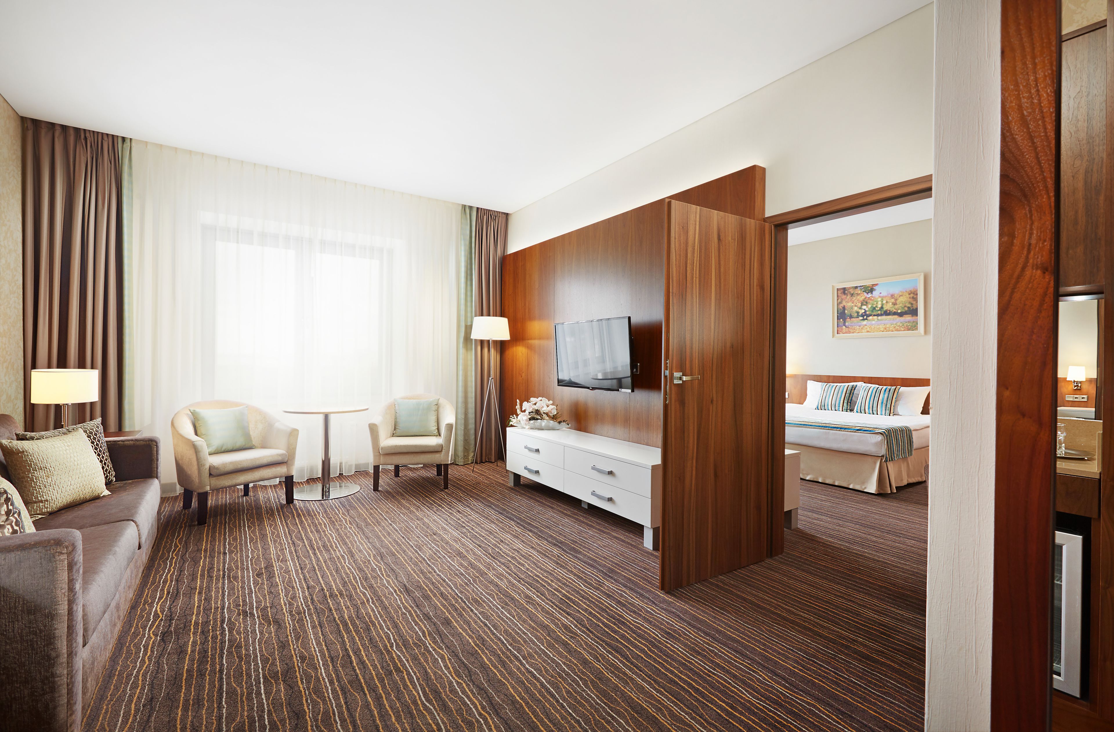 A hotel room with a brown carpet, a sofa, a chair, a TV, a bed, and a wooden wardrobe.