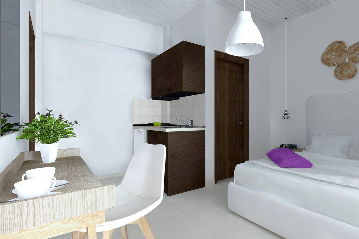 A modern bedroom with a bed, a desk with a chair, a small kitchenette, and a potted plant.